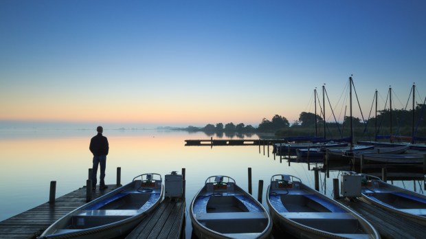 Man standing on a jetty in a marina during a foggy, autumn sunrise at a lake.