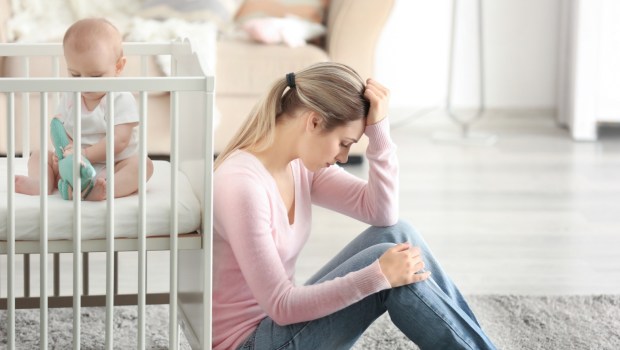 Zmęczona matka siedzi obok łóżeczka, w którym bawi się jej małe dziecko