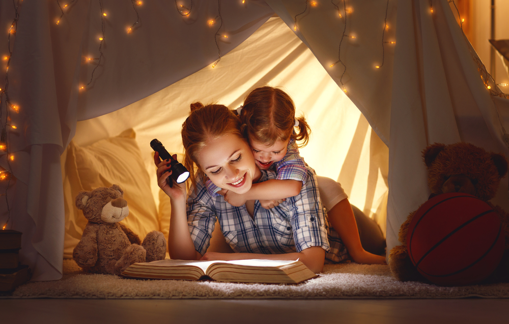 mama z córką czytają książkę pod namiotem z koca oświetlając ją latarką