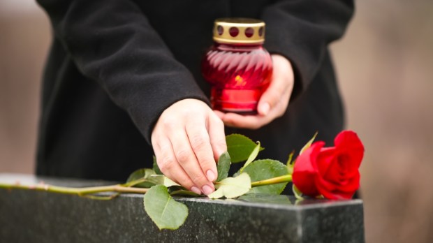 kobieta kładzie różę na nagrobku i trzyma w dłoni zapalony znicz