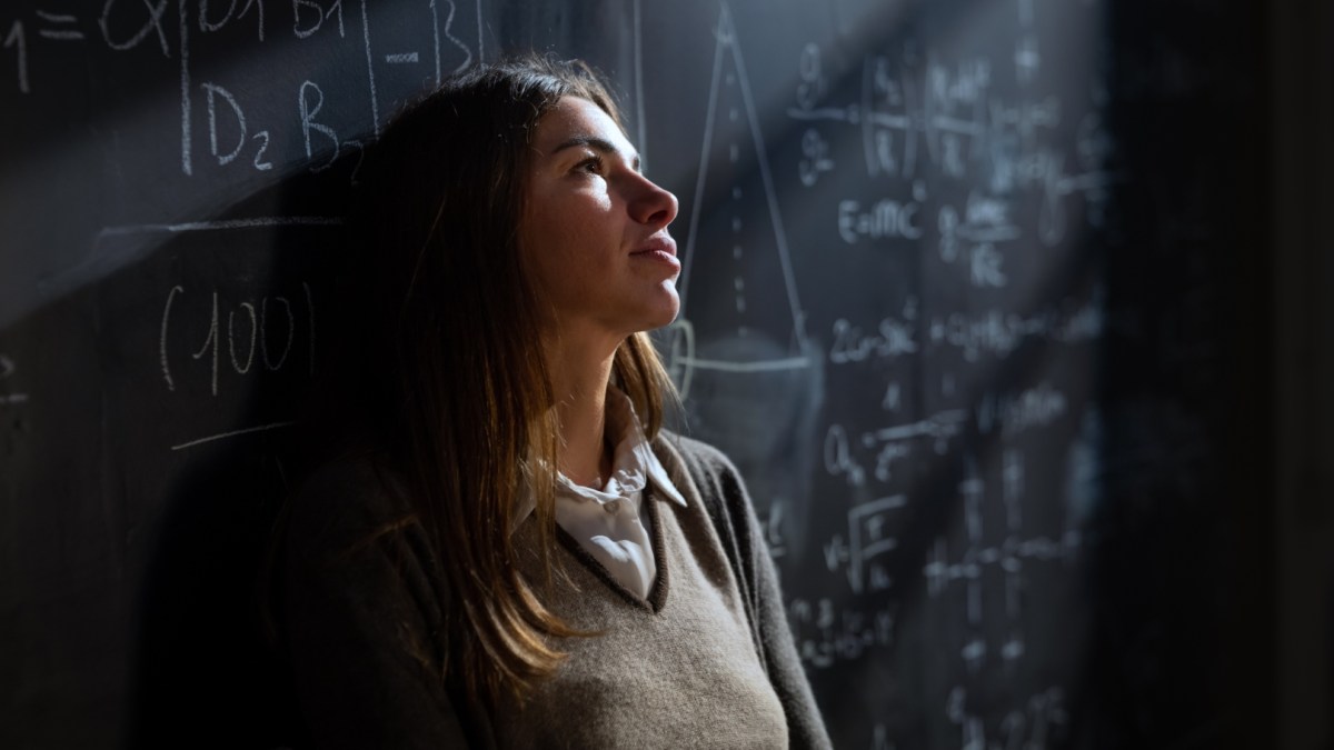Zamyślona studentka matematyki przed tablicą zapisaną wzorami matematycznymi i fizycznymi