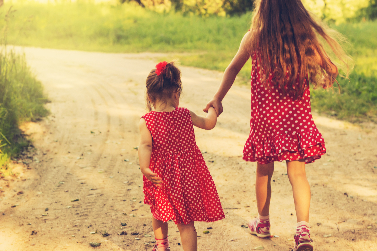 Dwie małe siostry w czerwonych sukienkach trzymają się za ręce i idą drogą