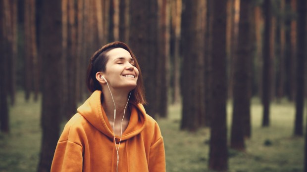 młoda dziewczyna ze słuchawkami na uszach stoi w lesie z zamkniętymi oczami i uśmiecha się