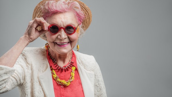 kolorowo ubrana uśmiechnięta starsza kobieta podtrzymuje dłonią okulary i patrzy w aparat