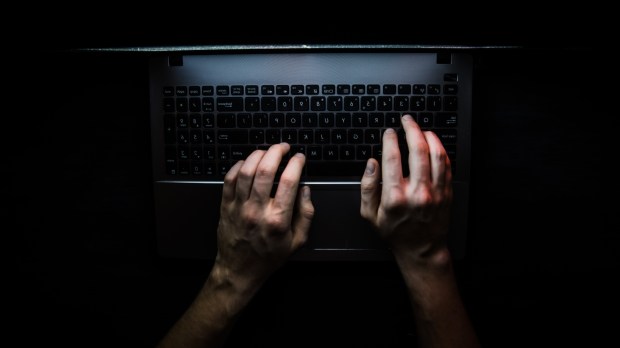 Dłonie internauty widziane z góry oparte na klawiaturze laptopa w zaciemnionym pokoju