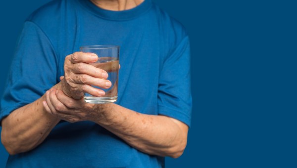 Chory na Parkinsona cżłowiek trzyma z niepewnością szklankę wody, pomagając sobie drugą ręką