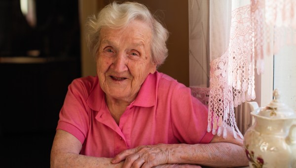 Uśmiechnięta staruszka siedzi przy stole koło okna w swojej kuchni