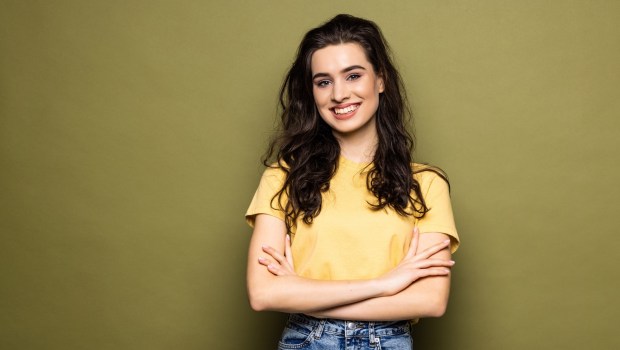 Young smiling attractive girl is posing in close pose with arms crossing on chest on khaki background looking at camera. Shy student, modest woman.