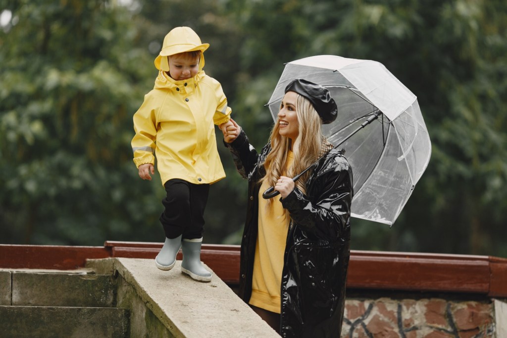 Dziecko w żółtym płaszczu przeciwdeszczowym wspina się po mokrych schodach trzymane za rękę przez mamę pod parasolem