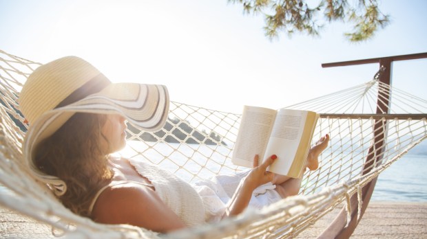 Kobieta w słomkowym kapeluszu leży w hamaku w pobliżu plaży w słoneczny letni dzień i czyta książkę