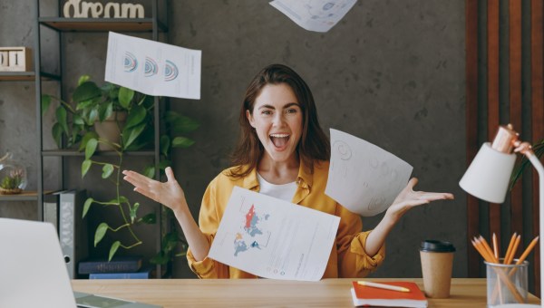 Kobieta siedzi za biurkiem i z radością wyrzuca kartki papieru w górę